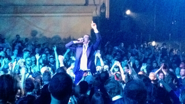 Singer Nick Cave standing on chairs in the middle of his audience, microphone to his mouth and left index finger pointed in the air.

